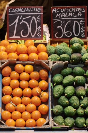 Cardboard boxes with oranges and avocados. They are displayed on the counter of a local business. There are two blackboards with text in Spanish that says