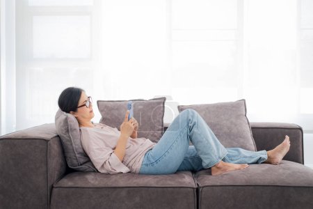 Foto de Feliz mujer asiática madura relajarse en cómodo sofá en casa mensajes de texto de mensajería en el teléfono inteligente, sonriente esposa uso de teléfono celular, navegar por Internet inalámbrico en gadget, compras en línea desde casa - Imagen libre de derechos
