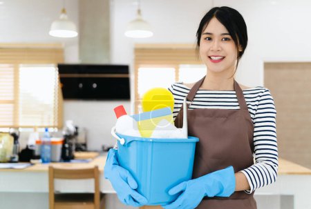 Foto de Servicio de limpieza. Primer plano del limpiador femenino sonriente, de pie en la cocina, sostiene cubo con detergentes y trapos, concepto de limpieza y limpieza - Imagen libre de derechos