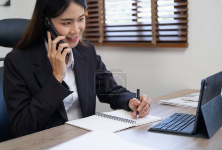 Foto de Feliz mujer de negocios llamando al teléfono móvil y tomando notas en un escritorio en la oficina - Imagen libre de derechos