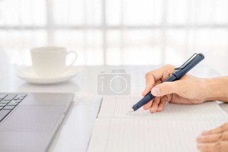 Foto de Mujer de negocios asiática en ropa casual escribiendo notas en bloc de notas mientras trabaja en el ordenador portátil, cerca de la imagen limpia frente a la ventana en luz suave. - Imagen libre de derechos