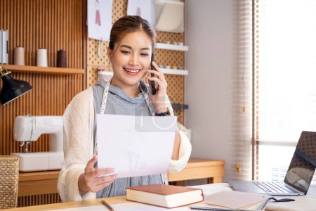 Photo for Dressmaking Sewing Service Worker Talking Phone. asian Woman Fashion Designer Holding Smartphone Writing Notes in Cozy Creative Design Studio or Tailor Shop. Beautiful Girl at Workplace - Royalty Free Image