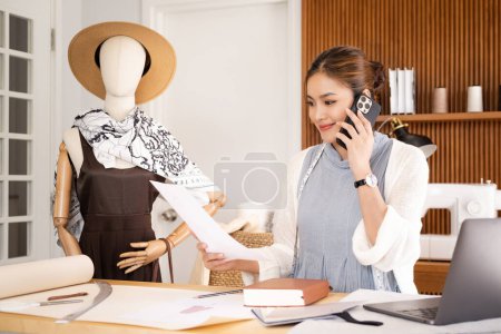 Photo for Smiling young asian fasion designer woman in blue white suit sitting in office of fashion designer and holds phone and paper talking with client . - Royalty Free Image
