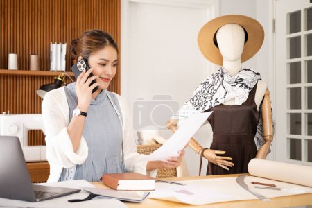 Photo for Dressmaking Sewing Service Worker Talking Phone. asian Woman Fashion Designer Holding Smartphone Writing Notes in Cozy Creative Design Studio or Tailor Shop. Beautiful Girl at Workplace - Royalty Free Image