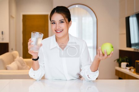 Foto de Asiática atractiva hembra sosteniendo un vaso de agua en la cocina. Joven hermosa mujer recoger manzana y beber mineral limpio natural en la taza después de despertar por la mañana para la salud y mirando a la cámara - Imagen libre de derechos