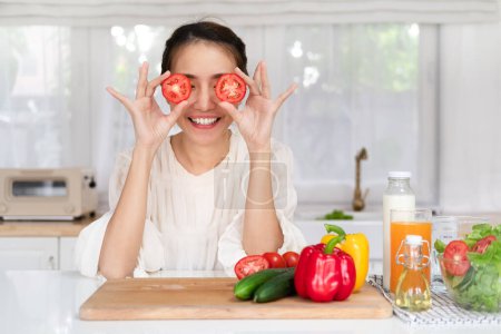 Foto de Feliz joven latina divirtiéndose y cubriendo los ojos con mitades de tomate, jugando en el interior de la cocina, mirando y sonriendo a la cámara mientras cocina la cena - Imagen libre de derechos