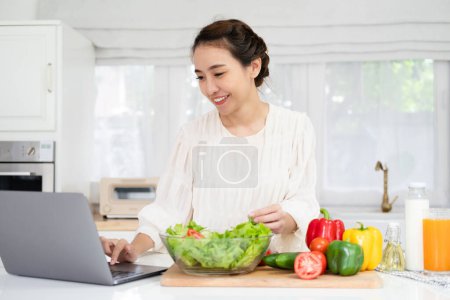 Foto de Tutorial de cocina en línea. Mujer joven asiática adolescente utilizando el ordenador portátil para bloguear bloguear en busca de receta mientras cocina comida vegetariana ensalada vegetal, hábitos alimenticios saludables. - Imagen libre de derechos