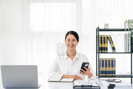 Foto de Atractiva morena mujer asiática usando el teléfono móvil, sonrisa y feliz mientras está sentado en el escritorio de trabajo en la oficina moderna. - Imagen libre de derechos