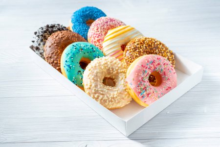 Sweet donuts of different bright colors lie in a box on a white table