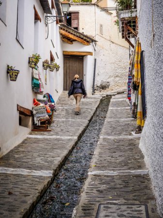 Junge blonde Frau, die eine Straße mit einem Wasserkanal in der Mitte der Straße im malerischen Dorf Pampaneira in den Alpujarras von Granada hinuntergeht