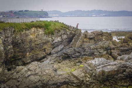 Téléchargez les photos : Jeune femme blonde dans une robe brune descendant les escaliers menant à une plage pleine de roches avec des algues collées à elle - en image libre de droit