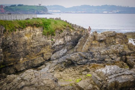 Téléchargez les photos : Jeune femme blonde dans une robe brune descendant les escaliers menant à une plage pleine de roches avec des algues collées à elle - en image libre de droit