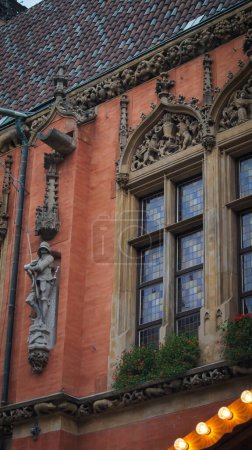 Foto de Un primer plano de una fachada del edificio histórico que muestra intrincados detalles arquitectónicos góticos. El exterior rojo-naranja está adornado con esculturas de piedra, incluyendo un caballero con armadura, figuras grotescas. - Imagen libre de derechos