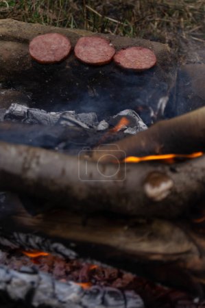 Smoky campfire grilling with savory sausages over embers. Rustic outdoor grilling, sausages sizzling on a wood fire. Sausages cook over a smoldering campfire, a taste of the wild. Open-flame cooking, hearty sausages on a natural campfire.