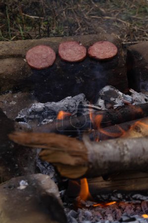 Tranches de saucisse grillées sur un feu de camp ouvert, cuisine extérieure. Saveurs fumées surgissent comme saucisses grésille sur un gril en pierre rustique. Barbecue du camping, saucisses salées sur une flamme de bois. Aventure culinaire en plein air avec saucisses rôties sur feu ouvert