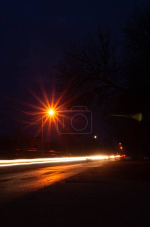 Evening falls as streetlights flicker on, casting light trails on the road. Urban twilight with street lamps glowing and cars painting streaks of light. Dusk settles on the city street, lights blur into luminous streams in the dark.
