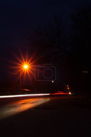 Téléchargez les photos : Un lampadaire rayonnant domine le crépuscule, avec des sentiers le long. La nuit se réveille comme des traînées de fléchettes lumineuses le long de la rue calme. Les lampadaires servent de sentinelles tandis que les véhicules esquissent des lignes de lumière dans l'obscurité. - en image libre de droit