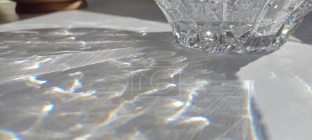 Sonnenbeschienene Kristallschale, die ein blendendes Lichtspektrum auf eine glatte Tischoberfläche wirft und die komplizierte Schönheit des gebrochenen Sonnenlichts in einer alltäglichen Umgebung einfängt.