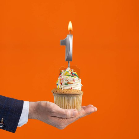 Téléchargez les photos : La main qui livre cupcake avec la bougie numéro 1 - Anniversaire sur fond orange - en image libre de droit