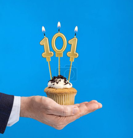 Téléchargez les photos : Main tenant un cupcake avec la bougie numéro 101 - Anniversaire sur fond bleu - en image libre de droit