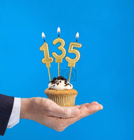 Hand hält einen Cupcake mit der Zahl 135 - Geburtstag auf blauem Hintergrund