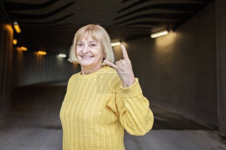 Téléchargez les photos : Femme de plus de 65 ans en pull jaune d'automne montre un geste de métal lourd. Elle est joyeuse et positive. Beau vieillissement et un regard optimiste sur la vie. Cornes, amour hard rock, signe. - en image libre de droit