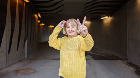 Téléchargez les photos : Femme de plus de 65 ans avec des lunettes en pull d'automne jaune montre geste de victoire. Elle est joyeuse et positive. Beau vieillissement et un regard optimiste sur la vie. Format de bannière - en image libre de droit