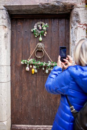 Foto de Una mujer mayor captura una encantadora puerta con temática de Pascua con una corona de huevo vibrante y una puerta de madera rústica en su teléfono inteligente, preservando los recuerdos de las vacaciones. - Imagen libre de derechos
