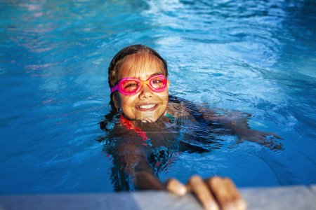 Foto de Una joven con una sonrisa radiante lleva gafas de color rosa mientras nada en una piscina azul brillante, encarnando la esencia de la alegría del verano. - Imagen libre de derechos