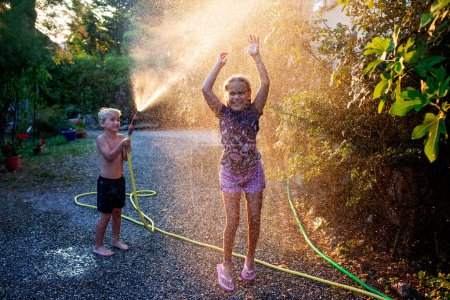 Fröhliche Kinder spielen im Sommerlicht mit Wasserschlauch, planschen und lachen in einem Hinterhof und schaffen Erinnerungen in der goldenen Stunde des Sonnenlichts.