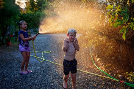 Foto de Los niños alegres juegan con la manguera de agua en la luz del verano, salpicando y riendo en un patio trasero, creando recuerdos en la luz del sol de la hora dorada. - Imagen libre de derechos
