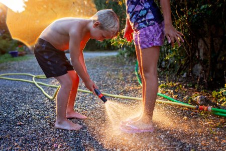 Siblings play with water in the sunset, brother spraying hose at sisters feet, sparking droplets in warm evening light.