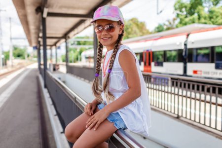 Foto de Chica sonriente con una gorra colorida y gafas de sol espera en una estación de tren, emocionado por una aventura de verano. - Imagen libre de derechos