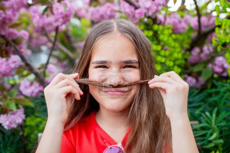 Young girl making a mustache with her hair, giggling in a garden full of pink blooms