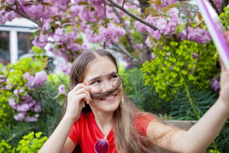 Foto de Chica que utiliza la tableta bajo el árbol en flor, compartir primavera belleza a través de videollamada, con bigote de pelo juguetón. - Imagen libre de derechos