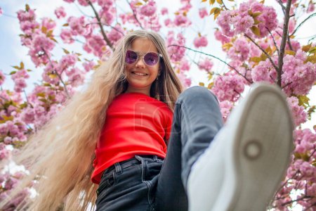 Foto de Una chica alegre se adelanta con una pose juguetona bajo un dosel de flores rosadas, sus gafas de sol reflejan el cielo primaveral. - Imagen libre de derechos