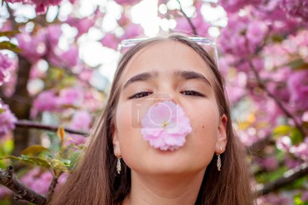 Foto de Una adolescente sostiene juguetonamente una flor de cerezo cerca de su nariz contra un telón de fondo de árboles florecientes. - Imagen libre de derechos