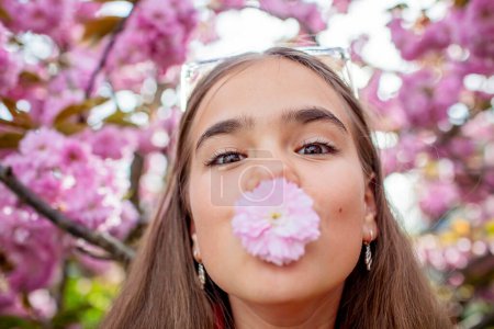 Foto de Una adolescente sostiene juguetonamente una flor de cerezo en su boca contra un telón de fondo de árboles florecientes. - Imagen libre de derechos