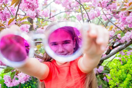 Foto de Una adolescente juguetona sostiene gafas de sol púrpura hasta la cámara, enmarcando su cara con vibrantes flores de cerezo. - Imagen libre de derechos
