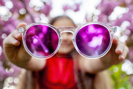 Foto de Una perspectiva creativa muestra a una chica sonriente sosteniendo gafas de sol púrpura con cerezos en flor en el fondo, se centran en las gafas - Imagen libre de derechos