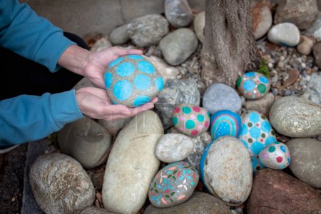 Hands of an elderly woman carefully holding a creatively painted rock, amongst a collection of various decorated stones