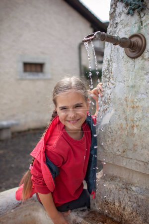 Foto de Una joven sonriente con una camisa roja juega con el agua que fluye de una antigua fuente del pueblo, disfrutando de un momento simple y alegre en un día soleado. - Imagen libre de derechos