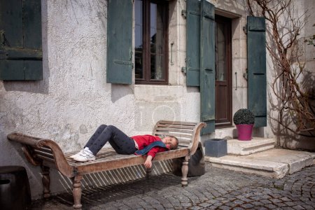 Foto de Una chica disfruta de un momento tranquilo en un viejo banco de madera junto a una casa tradicional con persianas verdes resistidas y una planta en maceta en la puerta. - Imagen libre de derechos