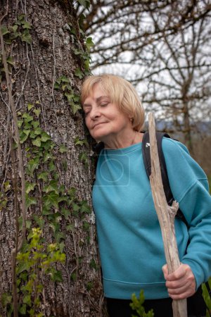 An elderly woman takes a moment of solace, leaning against a tree adorned with ivy, her eyes closed in peaceful reflection as she holds a walking stick.
