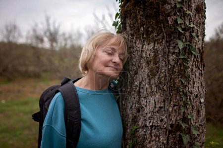 Une femme âgée avec une expression sereine repose contre un vieux tronc d'arbre, les yeux fermés, immergée dans la tranquillité de la nature lors d'une pause randonnée.