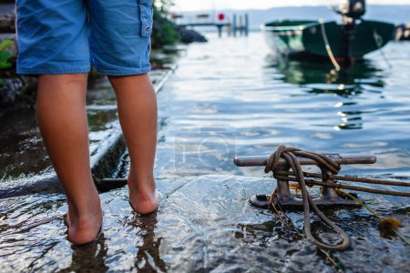 Ein Kind barfuß am Rande eines Sees, mit klarem Wasser und den Steinen. Verankertes Boot und ein mit einem Seil gebundener Metallsteg, Freizeit und Erkundung