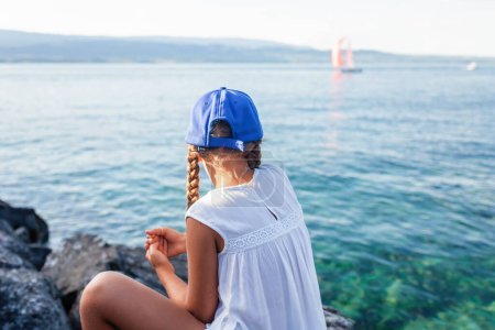 Foto de Una joven con una gorra de béisbol azul y un vestido blanco disfruta de un momento sereno junto al lago, viendo los veleros deslizarse por el agua.. - Imagen libre de derechos