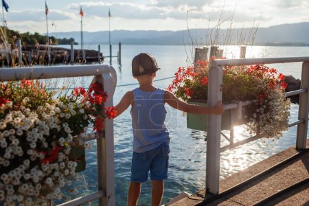 Foto de Un niño está parado en un muelle en un pequeño pueblo en el lago Leman, rodeado de floreros adornados, disfrutando del paisaje tranquilo y el toque suave de la puesta de sol en el agua. - Imagen libre de derechos