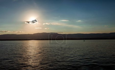 Foto de El sol se pone detrás de una nube sobre un lago tranquilo con montañas en la distancia, lanzando una escena tranquila. - Imagen libre de derechos