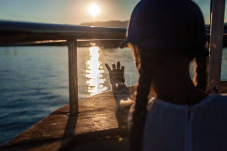 Foto de Niño en una gorra azul al atardecer en un barco, tocando el reflejo de la luz solar en el agua, evocando una sensación de asombro y la belleza de la naturaleza. - Imagen libre de derechos
